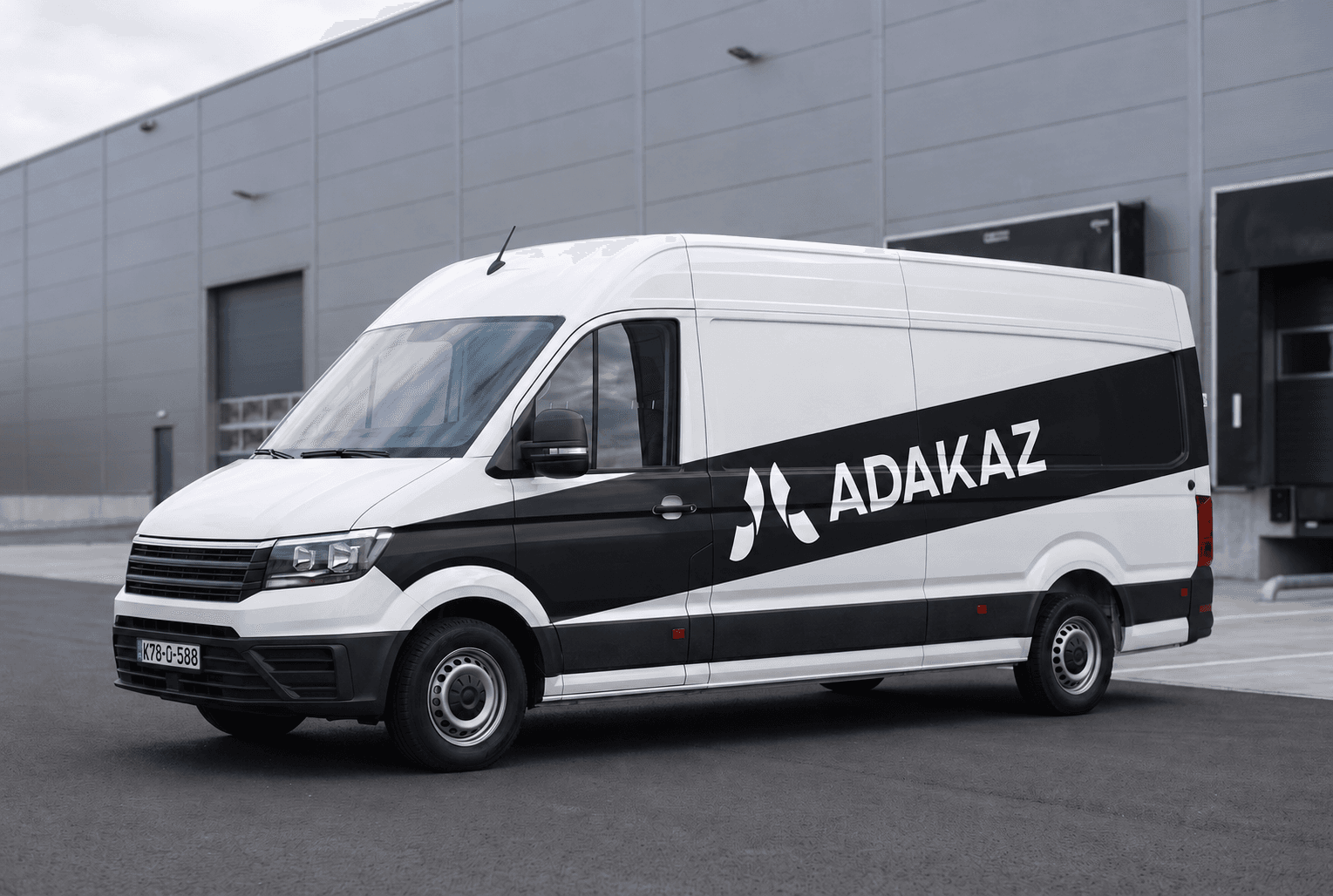 Bijelo dostavno vozilo s crnom dijagonalnom trakom i natpisom ADAKAZ, ispred logističkog skladišta.